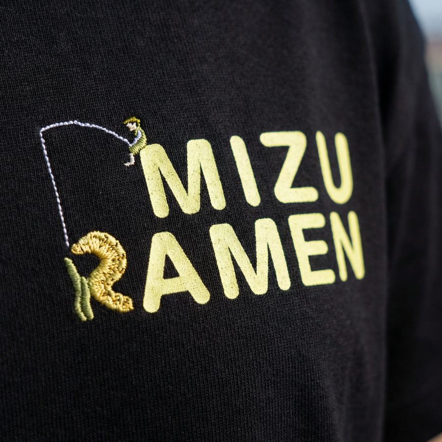 黒Tシャツの刺繍アップ。RAMENのRがウツボとワカメの刺繍に置き換わり、その上から釣り人が糸を垂らしている